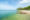 a long thin and pointed wooden boat on the shores of koh jum in thailand is parked in shallow and clear blue water and a stretch of white sea with a line of trees to the right