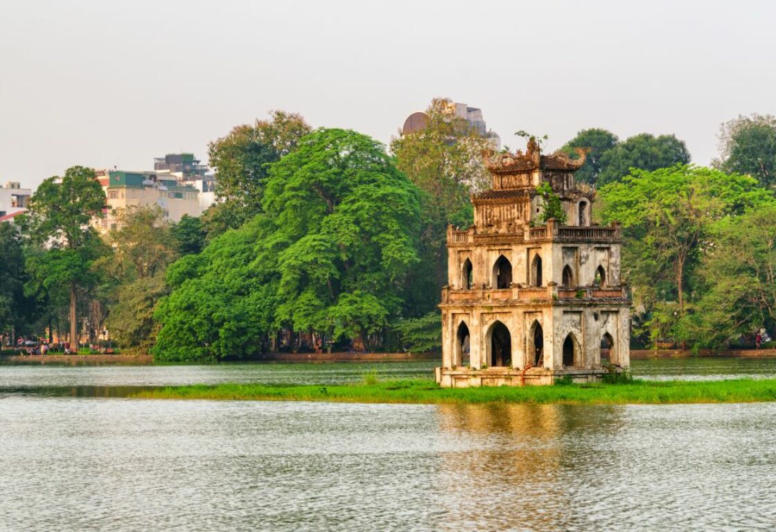Hoan Kiem Lake in Vietnam is a flat body of water and on the far side a line of trees and on the water a patch of grass has a small monument that has little arches all the way around the outside