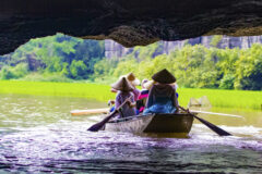 a group of 4 people sit in a boat going through a cave along the river and every person is wearing a conical hat and the cave emerges with a lush green bank on the side
