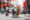 image of a street in hanoi with vietnamese flags hanging down and in the middle of the road a woman wearing a cone hat pushes a cart full of flowers and next to her a man rides a moped and a woman comes in another direction on a push bike