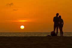 a man and a woman stand on the beach at sunset looking out to sea as the sun and sky glow orange