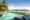 A turquoise infinity pool overlooking a calm bay with green rolling hills, palm trees, and blue umbrellas on the beachfront, with comfortable loungers in the foreground