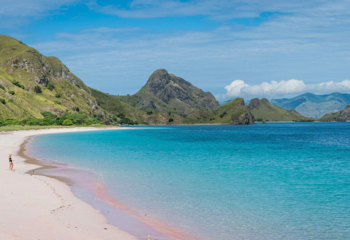 a woman stands on a stretch of perfect white sand in flores and the brilliant blue sea laps onto the shore turning the sand pink and cliffs and hills line the shore