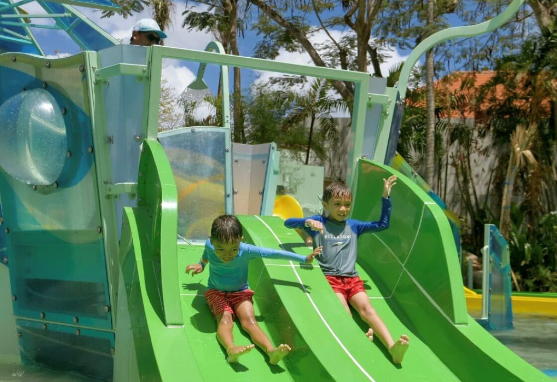 two young boys go down small green slides at a waterpark into a pool below