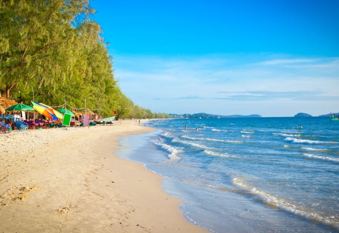 waves crashing onto sand in otres beach with a row of umbrellas near the palm trees