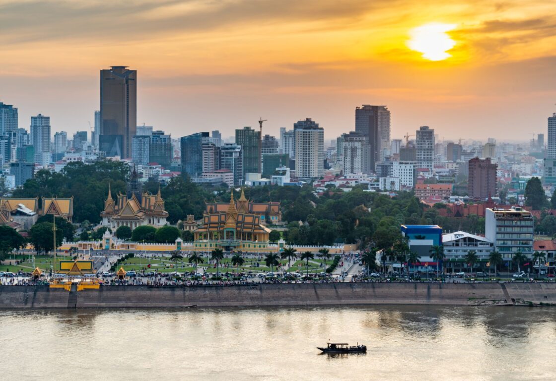 view across a river of the skyscrapers in phnom penh with temples in front and busy street and along the river at the bottom a single boat travels