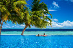 A couple relaxes at the edge of an infinity pool in the Maldives under the shade of palm trees, overlooking turquoise shallows and the deep blue ocean beyond, with a bright blue sky scattered with fluffy white clouds in the background.