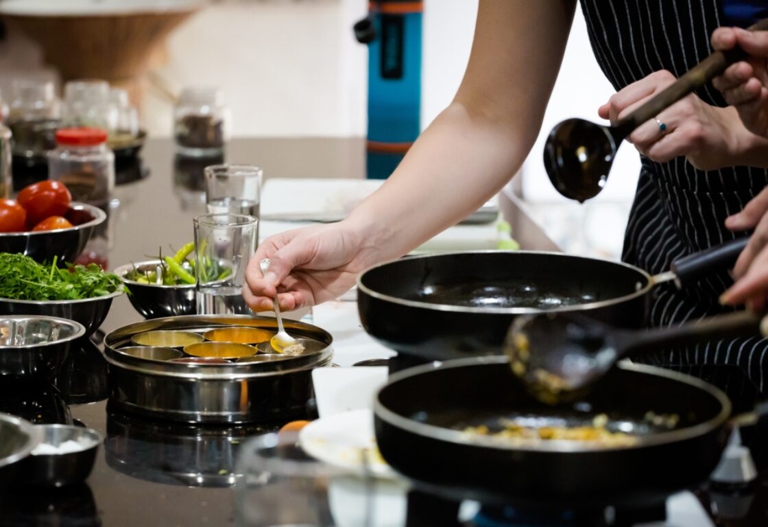 a cooking class in india with two black pans on a hob and a woman with a spoon scooping ingredients