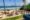View from a roof overlooking a sweep of the sea rolling onto the sand and on the sand is a large patio with tables and chairs dotted around the surface, and a few palm trees line the edge of the patio