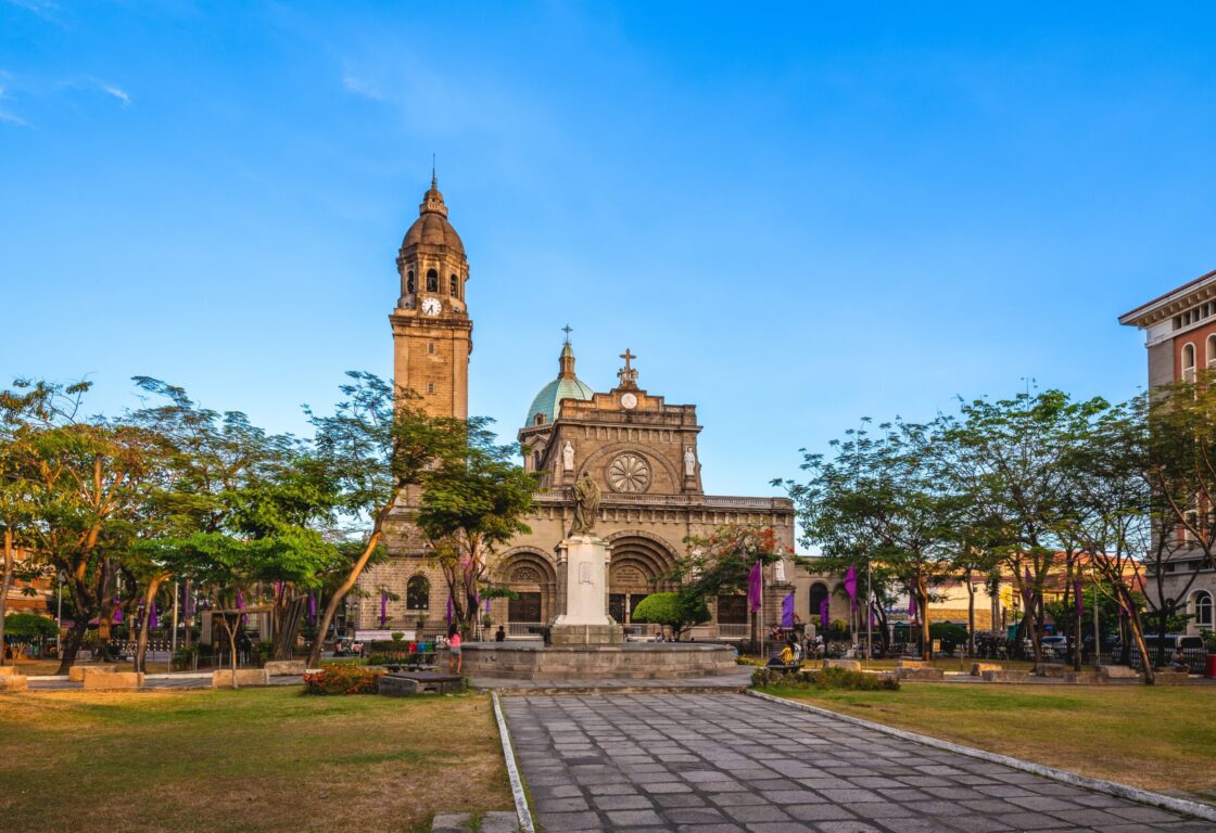 a shot of a cathedral with a tall clock tower on the left and in the distance, part of a dome can be seen and trees grown on either side