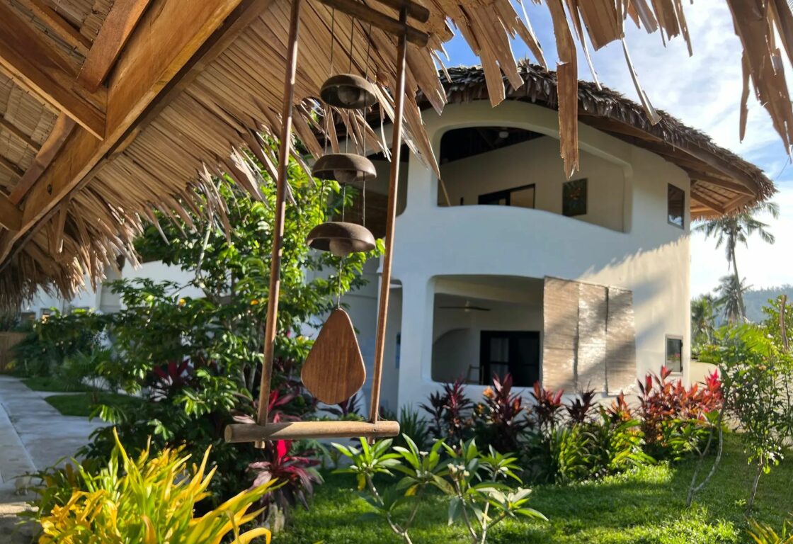 Parrots hotel in the philippines a white building with a curved edge and two large windows without glass on the right hand side and on the left a roof with thatched hangs down and a sun chime hangs from that roof