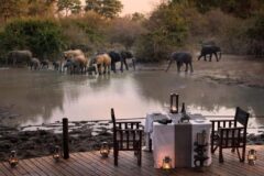 a view from a wooden terrace set wit a table for two with a table cloth and set for dinenr at sunset with elelphants drinking from a waterhole right in front of it
