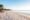 Tranquil beach scene at sunrise with palm trees, straw umbrellas, and lounge chairs on white sand. Calm sea stretches to the horizon under a clear sky.