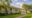 Charming house with a wraparound veranda, white columns, green roof, surrounded by lush gardens and trees, beneath a sunny blue sky with clouds.