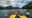 a family sits in yellow kayaks with two people in each, wearing blue coats and red life jackets holding oars out of the water and a guide in a blue kayak sits on the right hand side and the kayaks are on a flat lake with hills n either side and a cloudy sky