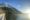 view across a flat lake at a snowy capped mountain and snow along the shore of the lake and the sun shines on the right hand side