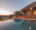 The main lodge and infinity pool at Garonga Safari Camp at dusk, with the lodge's dramatic curved thatched roof and warm interior lighting reflected in the still pool water, tall pillar candles on the pool deck, and a sweeping view over the African bushveld and dry riverbed at sunset.