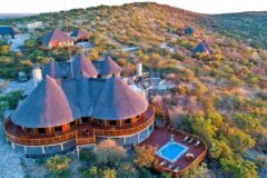an aerial view of a luxury safari lodge featuring a three dome roof, large viewing deck and terrace with a pool, small bungalows or safari tents around the property all located within a green trees and savannah area
