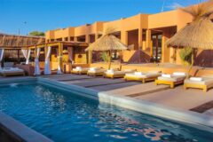 a pool area with white stone decking, wooden lounge chairs with white cushions, bamboo sun brollies, and individual cassitas behind it