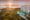 Sunset over a coastal hotel with a pool, surrounded by lush greenery. The sun casts a warm glow over the ocean, creating a tranquil atmosphere.