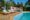 Poolside scene with three wooden lounge chairs with white cushions, shaded cabana with blue seating, lush palm trees, and bright blue sky. Relaxing tropical vibe.