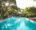a large pool with tiled patio around the edge and on the left hand side sun beds are lined up and a garden filled with trees and hedges are in the background and on the far side is a small wooden hut and on the left is a garden bench in the shade of the trees