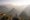 A solitary person stands on a rocky ledge with arms raised, overlooking a vast, misty mountain landscape with layered ridges and a winding river below in soft, golden light.
