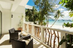 Villa Caemilla Boracay in the philippines two black armchairs on a terrace face a wall with pieces of wood in square gaps along it and beyond that there is white sand, palm trees and then a range of small boats on a calm blue sea