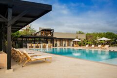 a pool area with a stone decking, lounge chairs and sun umbrellas with the main building behind it