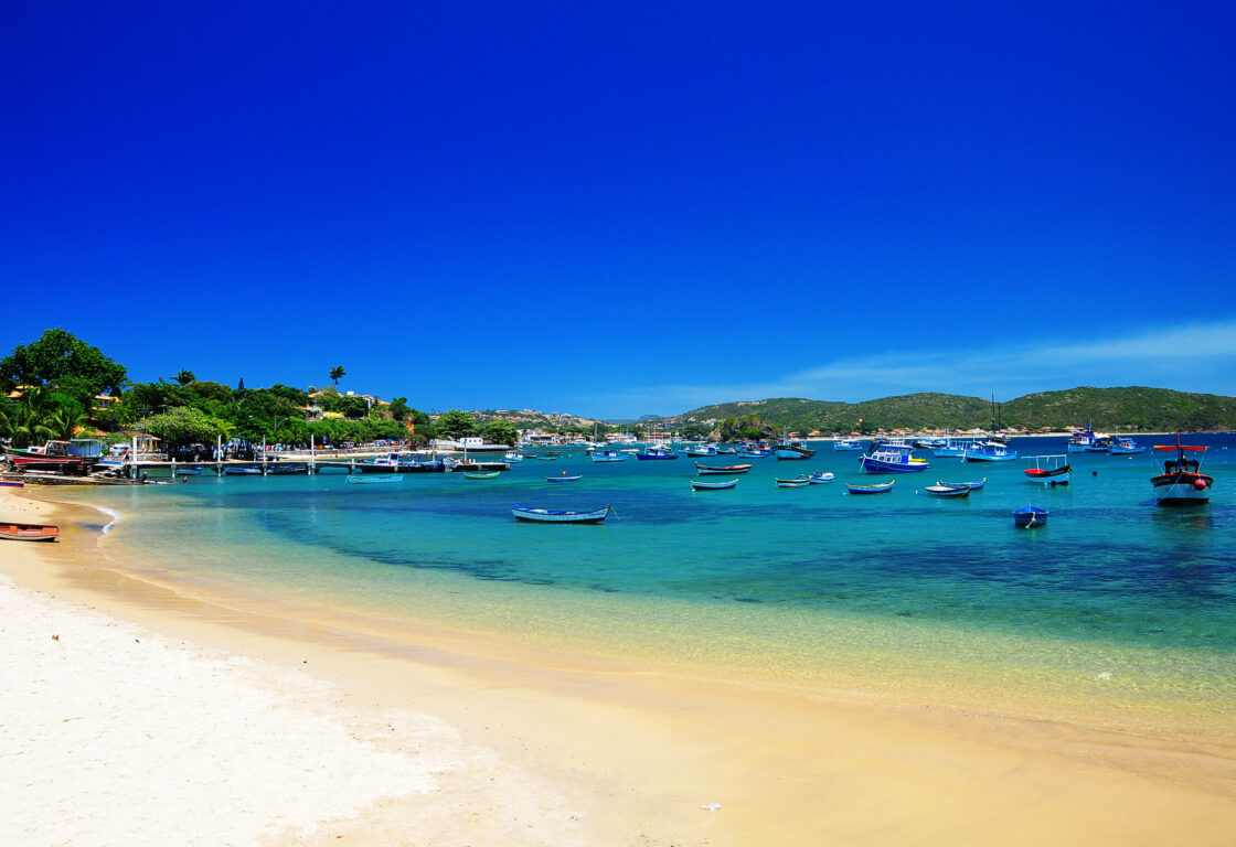 wide shot of white sandy beach with light blue coloured seawater. Dozens of small boats line the bay and in the distance are hills located on the other side of the bay