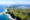 Aerial view of a coastal landscape with rolling green hills, winding roads, and scattered houses, bordered by a vibrant blue ocean with white waves. The serene setting conveys tranquility.