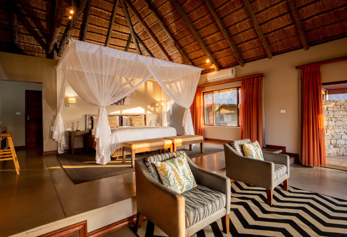 a large safari lodge bedroom with big bed and sheer white mosquito nets, stripy rug and two arm chairs with cushions, large windon and a wooden straw roof