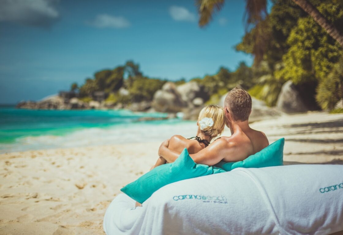 A couple sits on a beach, embraced on a white lounge with blue pillows, gazing at turquoise waves and lush greenery under a clear blue sky.