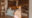 a close up double bed with white sheets and two large mustard coloured pillows on and a beige through and behind the wooden headboard is a wall made of flint stones and at the foot of the bed is a bench with cream coloured cushions
