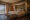 a large double bed has two small and fluffy cushions on and a piece of fabric is draped above the top of the bed that is up against a flint stone wall and on the left large glass doors reveal a view of a vineyard