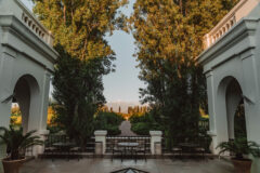 a large terrace with two square buildings that have archways on either side and in the middle of the terrace are black wrought iron table and chairs and the terrace overlooks a long path that is lined by low hedges and trees and the sun sets is casting a golden light across the scene