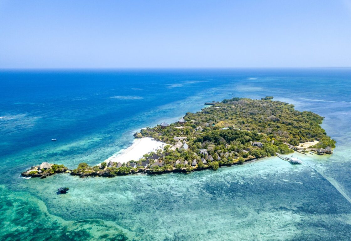 Aerial view of a small tropical island surrounded by clear turquoise waters, with white-sand beaches, thatched villas and coral reefs just offshore