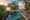 A view of a pool with stone flooring around it overlooking the river at sunset with trees throwing a shade