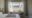 a bay window in the centre of a bedroom overlooks a field and mountains and floor to ceiling white curtains are open to the view, in front of the window are two armchairs and in the foreground of the image you can see the corner of a double bed with two brown blankets on