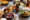 a table full of black bowl and plates that have a leafy salad on, tomato soup and other varieties of dishes and in the centre a small glass has some red wine in it