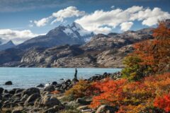 view of a man standing on a rock looking out to a lake with tall snow capped mountains in the background and on the right are trees covered in autumnal coloured leaves