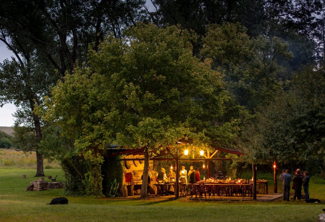 A cozy outdoor gathering under a lit gazebo, surrounded by trees, with people preparing food and enjoying each other's company at dusk.