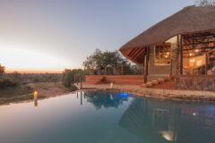 The main lodge and infinity pool at Garonga Safari Camp at dusk, with the lodge's dramatic curved thatched roof and warm interior lighting reflected in the still pool water, tall pillar candles on the pool deck, and a sweeping view over the African bushveld and dry riverbed at sunset.