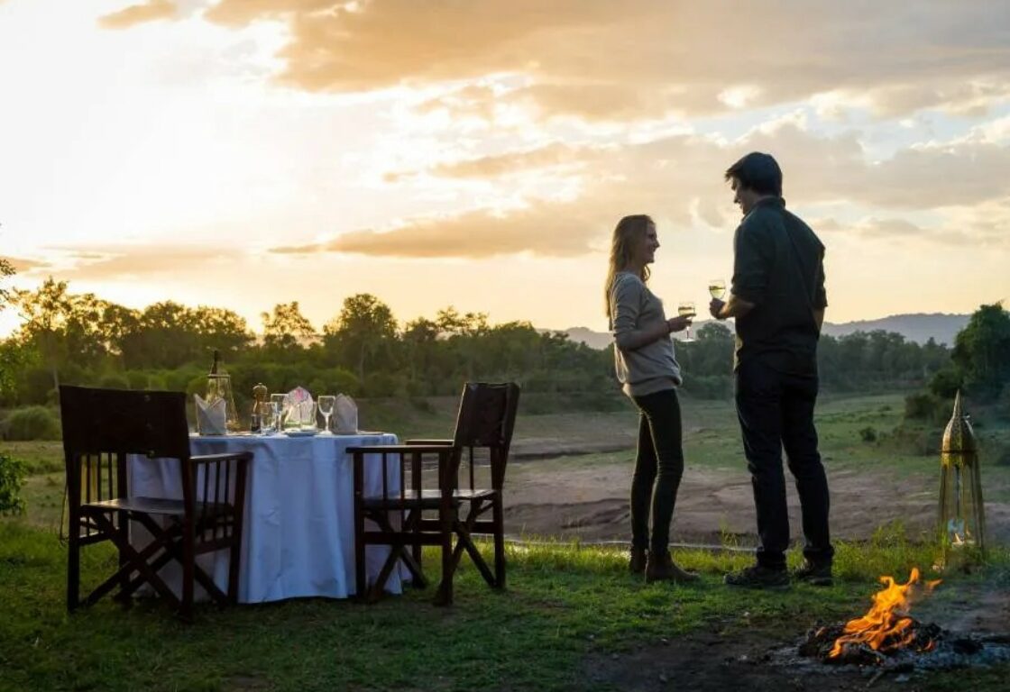 a couple standing together having a drink during a sunset with a table being set for a private dinner in the african bush