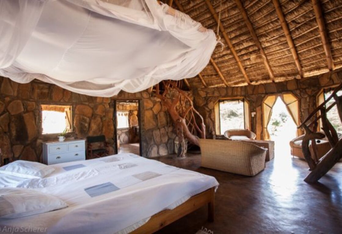 a view of a large bedroom inside of a tented safari lodge featuring a large bed with white bedding, seating sofa and big windows overlooking the african nature
