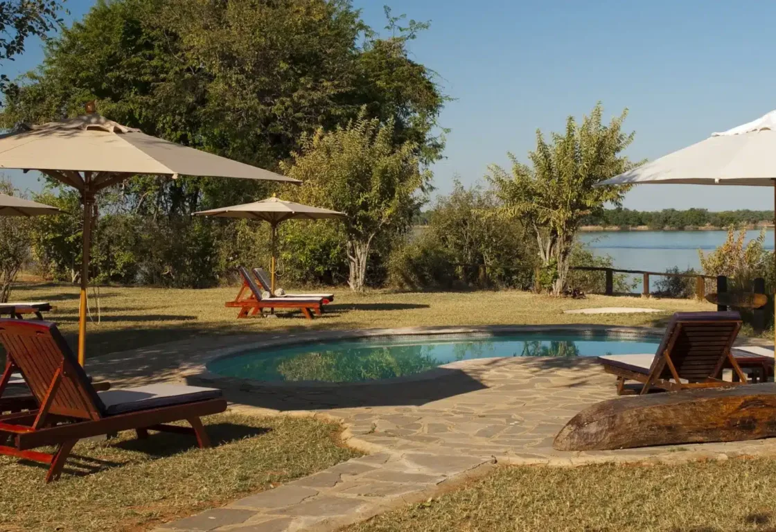 Poolside scene with wooden loungers and umbrellas on a stone patio. Overlooks a tranquil river, surrounded by lush greenery under a clear blue sky.