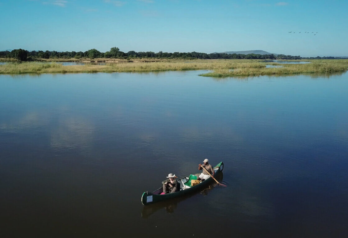 Aerial view of a canoe with two people on it, on a serenly calm river. In the distance is a shore, with green grassy marshland.