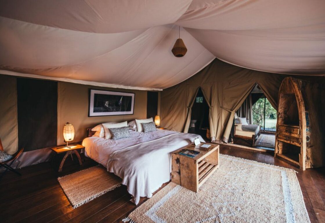 a safari tent with double bed featuring two dark coloured cushions, white rug and a small seater in front of it, small desk and a chair by the tent window