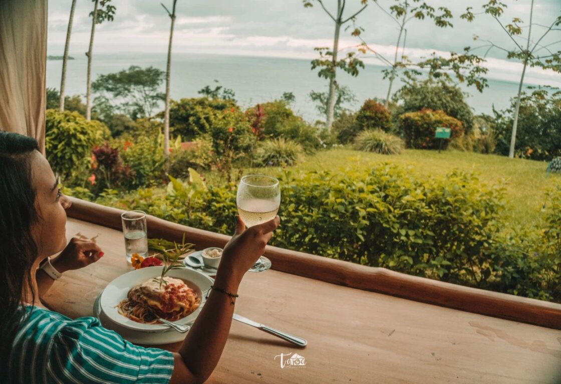 A person enjoys a meal overlooking a lush garden and ocean view, holding a glass of drink beside a plate of spaghetti.