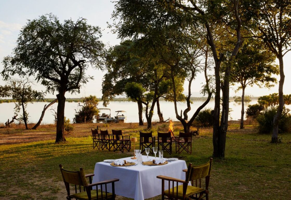 a table set on the river bank in Nyerere National Park in Tanzania, with boma fire and chairs in half circle overlooking the Rufiji River